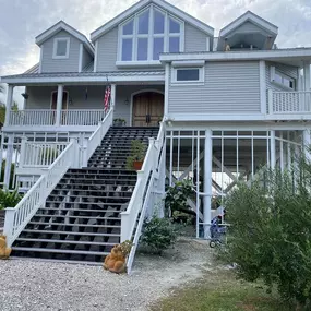 A large, elevated beach house with light gray siding, multiple balconies, and prominent staircases leading to the main entrance is shown.