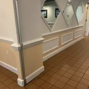 A hallway features light peach-colored lower walls with white wainscoting, diamond-shaped mirrors above, and brown tiled flooring leading towards a doorway.