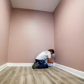 A person kneels in the corner of a small room with freshly painted light purple walls and gray wood-look flooring, seemingly touching up the baseboard.
