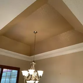 A low-angle shot shows a beige tray ceiling with textured paint, white crown molding, and a multi-light chandelier hanging from the center.
