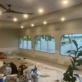 A bright room under renovation shows taped arched windows, unpainted walls, drop cloths, tools, and a potted plant in the foreground.