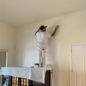 A person wearing a cap and white clothing stands on furniture covered with a white sheet, applying tape to a wall.