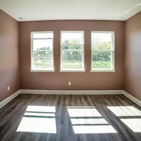 An empty room with freshly painted brown walls, white trim, and gray wood-look flooring is illuminated by sunlight streaming through three windows.