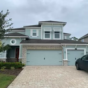 A two-story house with light blue siding, a dark roof, and a brick driveway is pictured on an overcast day. A dark SUV is parked.