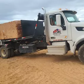 A white Ameri-Can roll-off truck transports a large, rectangular stone block on a dirt surface under a cloudy sky.