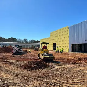 A construction site shows heavy machinery—a roller, skid steer, and bulldozer—working on a dirt lot next to a partially constructed building under a clear blue sky.