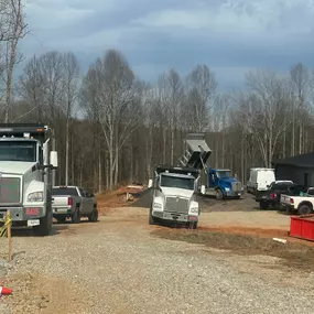 Several work trucks, including two Kenworth T880 dump trucks, and pickup trucks are parked on a gravel lot near bare trees and a building.