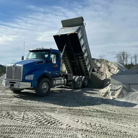 A blue 2025 Kenworth T880 dump truck is actively unloading gravel onto a large pile in a construction or material yard under a partly cloudy sky.