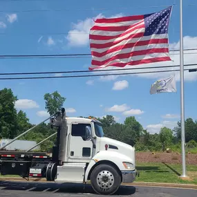 A white Kenworth T270 roll-off truck is parked near an American flag and a smaller, lighter-colored flag under a partly cloudy blue sky.