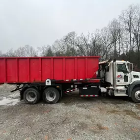 A white Western Star roll-off truck with a large red container is parked on a gravelly surface under an overcast sky near bare trees.