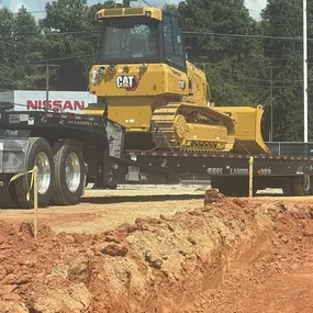 A yellow Caterpillar D3K2 bulldozer is secured on a flatbed trailer, ready for transport, in a construction zone with a Nissan dealership visible in the background.