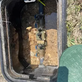 An open, rectangular black box in the ground reveals plumbing components. Pipes with valves and fittings are visible within the damp, earthen enclosure. Water has collected at the bottom. A partially visible green lid suggests this is an outdoor access point for plumbing maintenance or inspection.