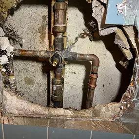 This close-up shows exposed copper plumbing pipes and fittings behind a damaged wall. The surrounding wall material is broken away, revealing the pipes connected to a central valve. Evidence of past water damage or repair attempts, such as discoloration and patching, is visible on the wall.
