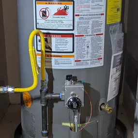 This image shows a Rheem 40-gallon gas water heater. A bright yellow gas line connects to the unit's control valve. Warning labels and an EnergyGuide sticker are visible on the side of the gray water heater. The appliance sits on a round metal pan, likely for leak protection.