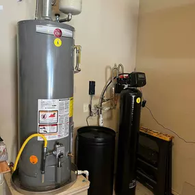 This well-lit utility space features a gray Rheem gas water heater with a yellow gas line and associated plumbing. Beside it sits a black water softener tank and a black brine tank. The setup suggests a home plumbing system providing hot water and water treatment.