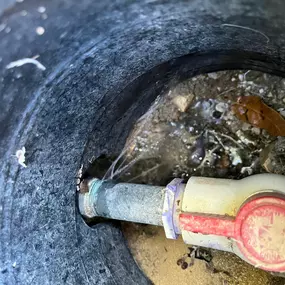 Two open access boxes in the ground reveal plumbing infrastructure. The top oval box shows a pipe and valve. The lower rectangular box contains a flow meter with blue and white fittings, surrounded by gravel. This suggests an irrigation or water management system.