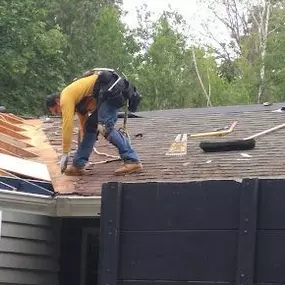 The image shows a construction worker in a yellow long-sleeved shirt, blue jeans, and safety harness working on a roof. Part of the old roof has been removed, revealing wooden beams. Tools and a trash can are visible on the remaining shingles.