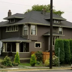 The image shows a large, two-story Craftsman-style house with dark brown siding, a dark grey roof, and light-colored trim around the windows and eaves. Mature landscaping surrounds the property, and a car is parked on the street in front.