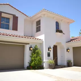 A modern Mediterranean-style white house features a terracotta roof, two beige garage doors, and a landscaped entryway with plants under a clear blue sky.