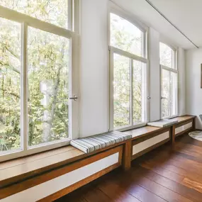 A room features large windows overlooking a tree, with wooden benches, a gray chair, and a black canvas on the wall.