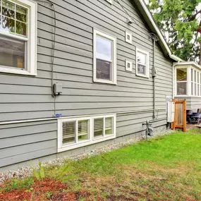 The image shows a house with gray siding, several windows, a small bay window extension, and a green lawn in front.