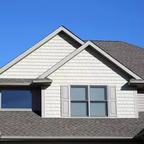 The upper portion of a house shows a roof with gray shingles, light-colored siding in the gables, and several windows against a clear blue sky.