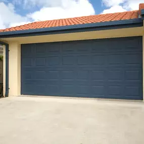 A blue, sectional garage door is attached to a beige garage with a red tiled roof and a concrete driveway.