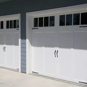 Two white garage doors with windows and decorative black hardware are attached to a building with blue siding.