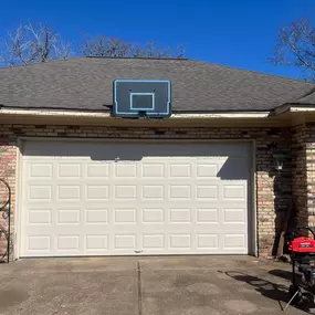 A basketball hoop is mounted above a closed, white garage door on a brick building under a clear blue sky. Bare tree branches are visible above the roofline.