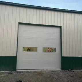 A white garage door with two small windows is set in a light-colored metal building. Green trim accents the top and bottom of the building's exterior.