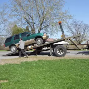 A green SUV is being loaded onto a flatbed tow truck by a person on a grassy roadside under a bright blue sky.