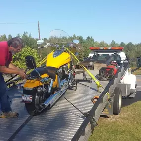 A person, likely of European descent, is securing a yellow motorcycle onto a flatbed tow truck on a grassy roadside.