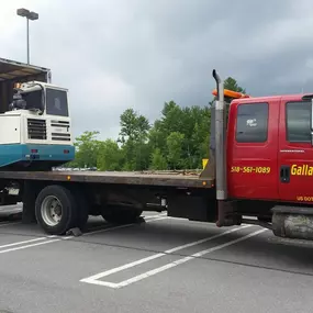 A red Gallagher's Towing truck is loading a blue and white piece of equipment from a white trailer in a parking lot.