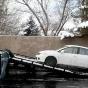 A white car is being loaded onto a black flatbed tow truck on a snowy street, with snow-covered trees in the background.