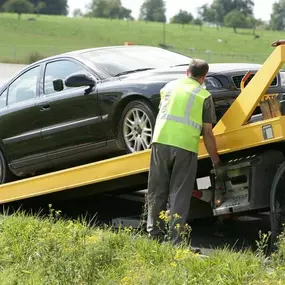 A person in a safety vest is operating a yellow tow truck to load a dark sedan on the side of a highway.