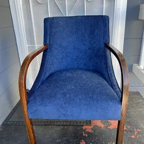 A blue upholstered armchair with curved wooden armrests stands on a porch with a white door and decorative metalwork in the background.
