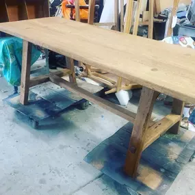 A long, light-brown wooden table with a trestle base is shown in a workshop setting, likely undergoing restoration.
