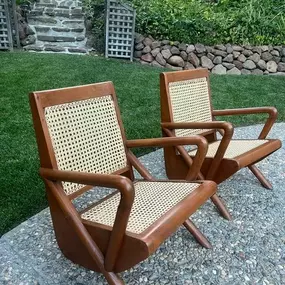 Two Mulholland caned armchairs with wooden frames and woven seats are positioned outdoors on a stone surface, with greenery and stone steps in the background.