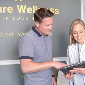 In the foreground, a smiling female provider in a white coat (likely you, Julia) is holding a tablet and reviewing information with a male patient in casual attire. Both are engaged and appear collaborative, suggesting a patient-centered, approachable atmosphere.

The background wall features the Venture Wellness logo in gold lettering, with the tagline “Elevate Your Health.” This adds brand presence and reinforces professionalism.

The setting is bright, modern, and welcoming, with light walls,