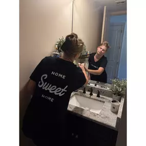 This image beautifully showcases a Breathe Maids of Orlando cleaning professional meticulously wiping down a bathroom mirror and sink area. She is wearing a branded black t-shirt with the words 