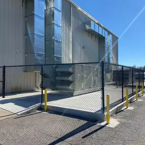 A black chain-link fence, accented with yellow posts, encloses industrial equipment outside a large metal building. Silver ductwork rises vertically along the building's exterior.