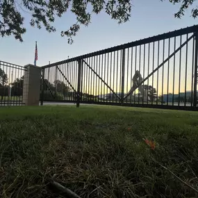 A closed black metal cantilever sliding gate stands on a green lawn under a dusky sky with hints of sunrise or sunset. A brick pillar supports the gate, and a US flag flies nearby. Trees and buildings are visible in the blurred background.