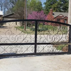 A decorative black wrought iron automatic gate is partially open, revealing a driveway leading to houses. The gate is flanked by light-colored stone pillars and a black metal fence. A gate opener is visible on the right side, and a warning sign is posted on the left.