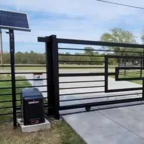 A black 16' Comunello Cantilever gate with a unique geometric design is installed on a concrete surface. A solar panel is mounted on a pole near the gate operator.