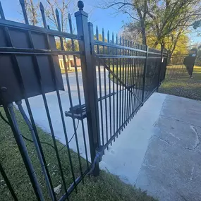 A black ornamental metal fence and automatic gate are partially open on a sunny day. The gate operator is visible, along with wiring. The fence runs along a concrete surface and grassy area, with trees and a street with a car in the background.
