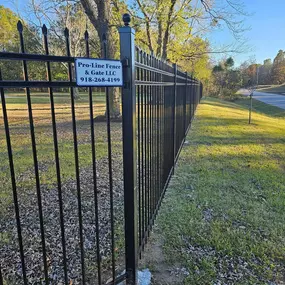 A black ornamental metal fence and automatic gate are partially open on a sunny day. The gate operator is visible, along with wiring. The fence runs along a concrete surface and grassy area.