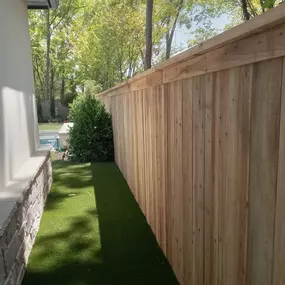 A light-brown wooden fence runs along a green, grassy area, with trees and greenery visible in the background. The fence is newly installed and contrasts with the green lawn.