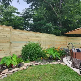 A backyard scene features a light-colored horizontal wooden fence enclosing a garden with various green plants, including some with pink coneflower-like blooms. A patio area with a wicker couch, pillows, and a tan umbrella is visible in the corner.