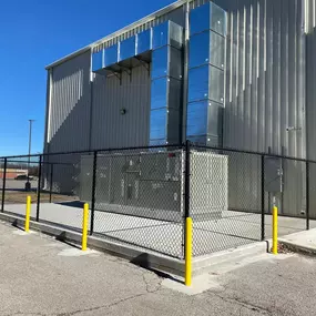 A black chain-link fence, accented with yellow posts, encloses industrial equipment outside a large metal building. Silver ductwork rises vertically along the building's exterior.