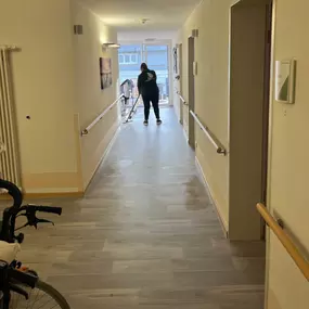 Eine Mitarbeiterin von Fein Gebäude-Service reinigt sorgfältig den Boden in einem hellen Flur mit Haltegriffen – eine Umgebung, die auf barrierefreie Pflegeeinrichtungen hinweist.